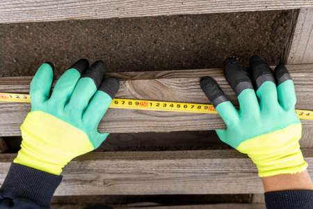 Gloved hands measure a wooden board with a tape measure. Construction and repair