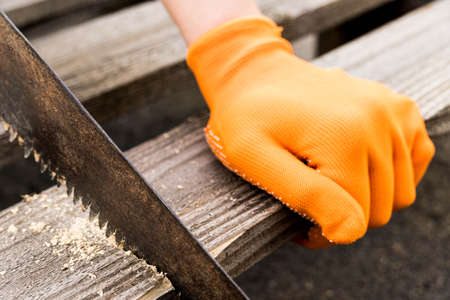 A hand in an orange glove is sawing a board with an old rusty hand saw. Construction and renovation conceptの写真素材