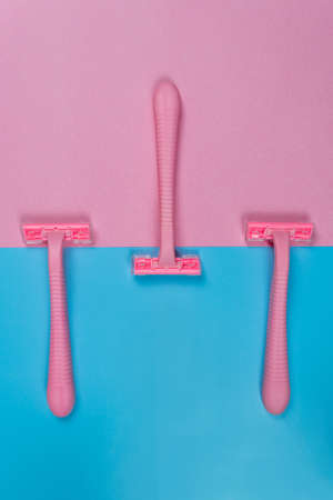 An overhead view of three new disposable pink razors lie on a blue-pink background with copy space....