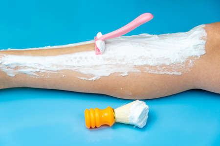 The procedure for hair removal of the legs with a pink disposable razor on a blue background, next to it is an orange shaving brush soaked in shaving foam. Personal hygiene of a womanの写真素材