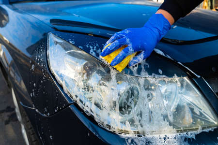 A female hand in a blue glove washes the headlight of a car with a yellow foam sponge. Car care and washingの写真素材