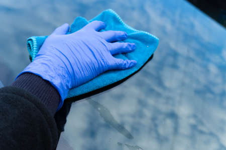 A woman's hand in a blue glove rubs the glass of a car in which the clouds are reflected. Car...
