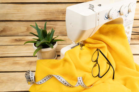 White sewing machine with yellow cloth, centimeter tape and glasses, in the background a flower in...