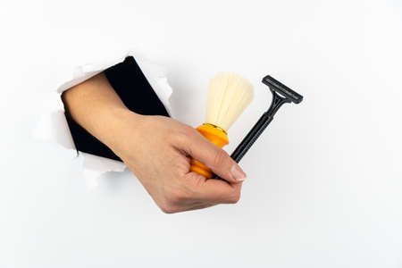 Shaving brush and razor in a woman's hand. The hand goes through a hole in the white paper wall. ... Means for removing hair from the skin. Free space for textの写真素材