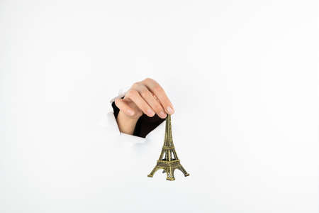 A female hand holds the eiffel tower statuette by the top on a white background. Hand goes through a hole in a white wall Free space for textの写真素材
