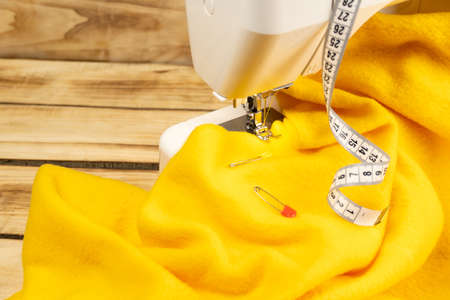 A piece of yellow fabric on a sewing machine and a centimeter on a wooden background. The fabric under the sewing machine foot there are two pins on it starts the sewing process. Free space for textの写真素材