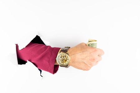 A man hand in a burgundy sleeve of a jacket with a watch sticking out of a hole in a white wall with a dollar. Business and time conceptの写真素材
