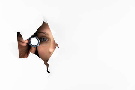 A woman in her thirties shines a flashlight through a hole in a white wall. Free space for textの写真素材