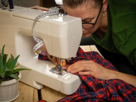 A woman of about thirty in glasses sews on a sewing machine on a wooden background. Sewing and...