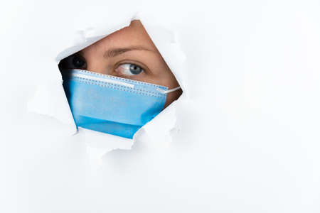 A woman wearing a medical mask looks through a hole in a white paper wall. Fear of coronavirus disease. Free space for text.の写真素材