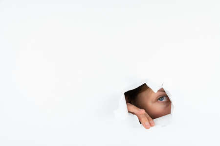 A woman peeks through a hole in a white paper wall with one eye. Free space for textの写真素材