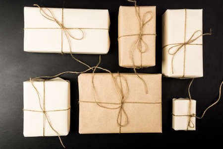 Top view of gifts on black background. Gifts are packed in white paper and tied with a rope from a...