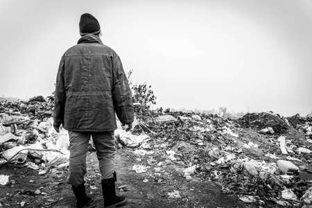 A woman in old clothes stands against the background of a landfill. Free space for text. Black and...