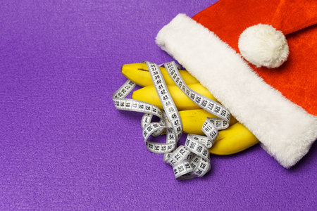 Yellow bananas and a centimeter tape on a purple background wearing a santa claus hat. Healthy food concept for Christmas holidaysの写真素材