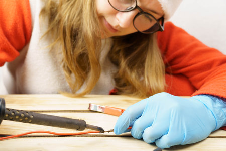 The woman solders the wires with a soldering iron. Female hands in rubber gloves hold a soldering iron over the wires. Soldering, maintenance and repair work in the workshopの写真素材