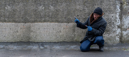 A filthy man, leaning on one knee, points his forefingers at a gray wall. Free space for textの写真素材