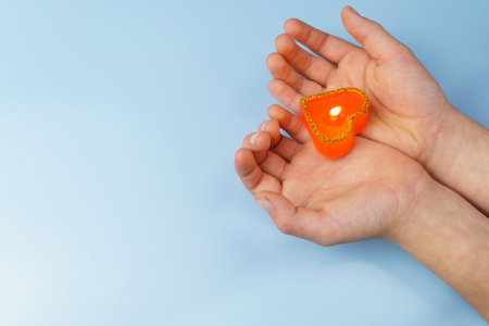 Burning red candle in the shape of a heart in children's hands on a blue background. Valentine's Day card.の写真素材