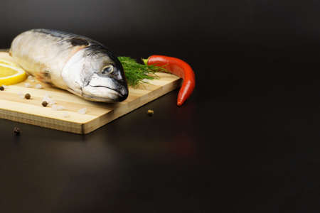 Raw mackerel, chili peppers, fresh dill, black pepper and lemon on a brown wooden cutting board, over black background with copy space. Seafood, healthy food and cook at home concept.の写真素材