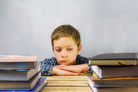 A tired frustrated schoolboy sits among a stack of books on a wooden table. World Book Day,...