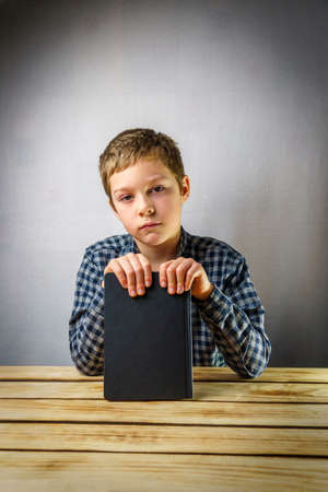 Sad boy from 7 to 9 years old, holding a book in his hands, on a gray background at a wooden table. Learning and education concept.の写真素材