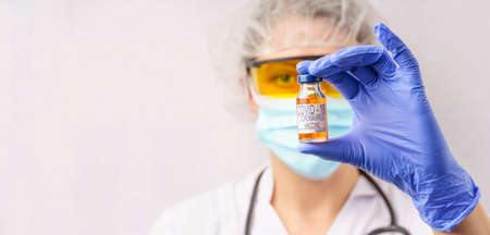 Female doctor wearing gloves, a protective mask and a bottle labeled COVID-19 CORONAVIRUS VACCINE. Gray background, free space for textの写真素材