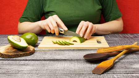 The girl cuts a ripe green avocado with a knife. Cooking. The fruits are cut on a wooden cutting board.の写真素材