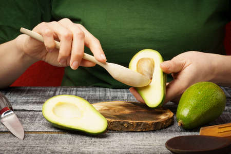 The girl cleans the avocado with a wooden spoon. Young woman with fresh avocado at the table