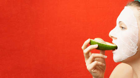 young woman in a tissue cosmetic mask biting a ripe avocado. girl on red wall background with copy spaceの写真素材