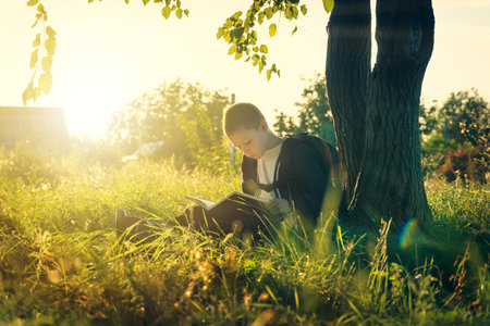boy sitting under a tree reads a book. school-age child learns in nature. the sun illuminates it. bannerの写真素材