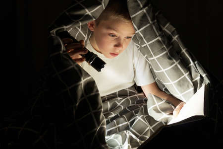 a boy under a blanket reads a book in the dark with a flashlight.
