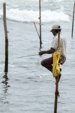 Stilt fisherman with his wooden rod facing side to the camera with his yellow pocket on the pole,...