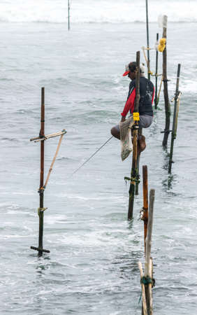 Stilt fisherman with his wooden rod facing side with his pocket on the pole to fill with fish,...