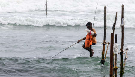 Weligama, Southern Province / Sri Lanka - 07 26 2020: Stilt fishermen with his wooden rod facing side to the camera with his orange pocket, fishing in a traditional unique method in Sri Lankan cultureのeditorial素材