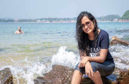 Cute south Asian girl with spectacle and short jeans sunbathing, sitting in a rock in jungle beach, Galle, ocean waves crashing behind her as her hair his wet in salt water,のeditorial素材