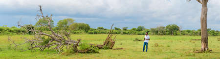 Hambantota, Sri Lanka - 04 16 2021: Fallen tree and a young boy panoramic view of the Lunugamvehera National Park, dark gloomy rain clouds gathering in the backgrounds,のeditorial素材