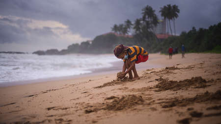Small kid wearing a face mask and a hat, Digging up beach and collect sand in both hands.の写真素材