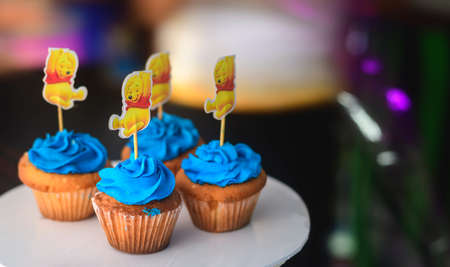 Four cupcakes with creamy swirled icing toppings in a cake stand in an indoor party celebration. Winnie the pooh cartoon character in a stick on top of the delicious tasty cupcakes.の写真素材