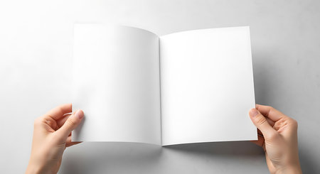 A top down view of two hands holding an open blank white brochure or magazine against a light background.の素材