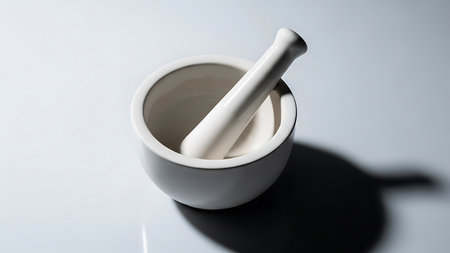 A clean white ceramic mortar and pestle rests on a smooth, light gray surface, casting a deep, sharp shadow.の素材