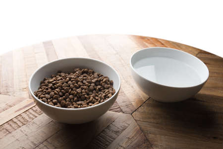 cat food dishes - two white bowls of cat dry food and water standing on a wooden table in front of white backgroundの写真素材