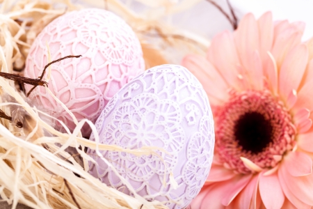 Beautiful pastel coloured Easter eggs in handcrafted intricate crocheted covers with a fresh pink Gerbera daisy in a clean straw nest for a traditional symbolic Easter celebrationの写真素材