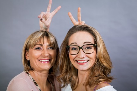 Playful beautiful young mother and her teenage daughter posing together with the young girl peeking out to the side with a happy grin, isolated on a grey studio backgroundの写真素材