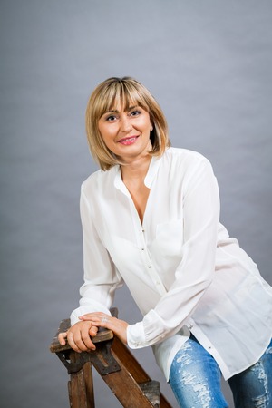 Smiling confident middle-aged blond woman in a fresh white blouse standing with her hand on her hip smiling at the camera, on greyの写真素材