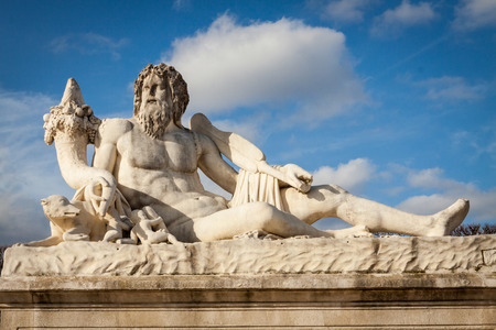 Bird perched on an ancient stone statue of a seated god or deity with a long curly beard and hair in Paris, France, possibly part of a stone fountain, viewed low angle against a cloudy blue skyの写真素材