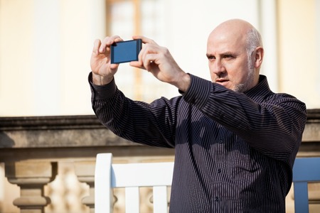 Attractive bald middle-aged man sitting on a wooden bench in an urban setting taking a photograph with his mobile phoneの写真素材