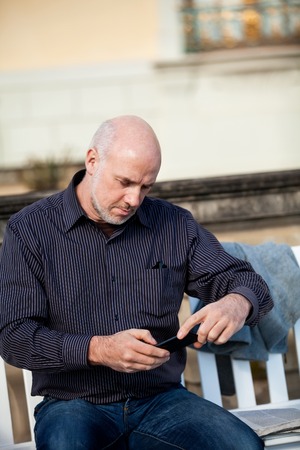 Attractive middle-aged man sitting on a wooden bench in an urban environment checking a photo on his mobile phoneの写真素材