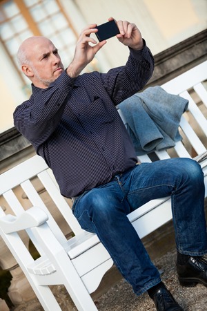 Attractive bald middle-aged man sitting on a wooden bench in an urban setting taking a photograph with his mobile phoneの写真素材
