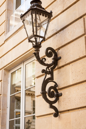 Old ornate wrought iron lamp with a scrolled frame and rectangular glass lantern mounted on a wooden building exteriorの写真素材