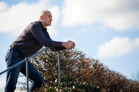 View from below of a thoughtful middle-aged man sitting on a flight of steps staring into the distance with a serious expression against a blue skyの写真素材