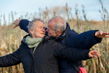 Attractive elderly couple in warm clothing standing clue together with outstretched arms, closed eyes and laughing smile against a blue sky embracing and celebrating the sunの写真素材